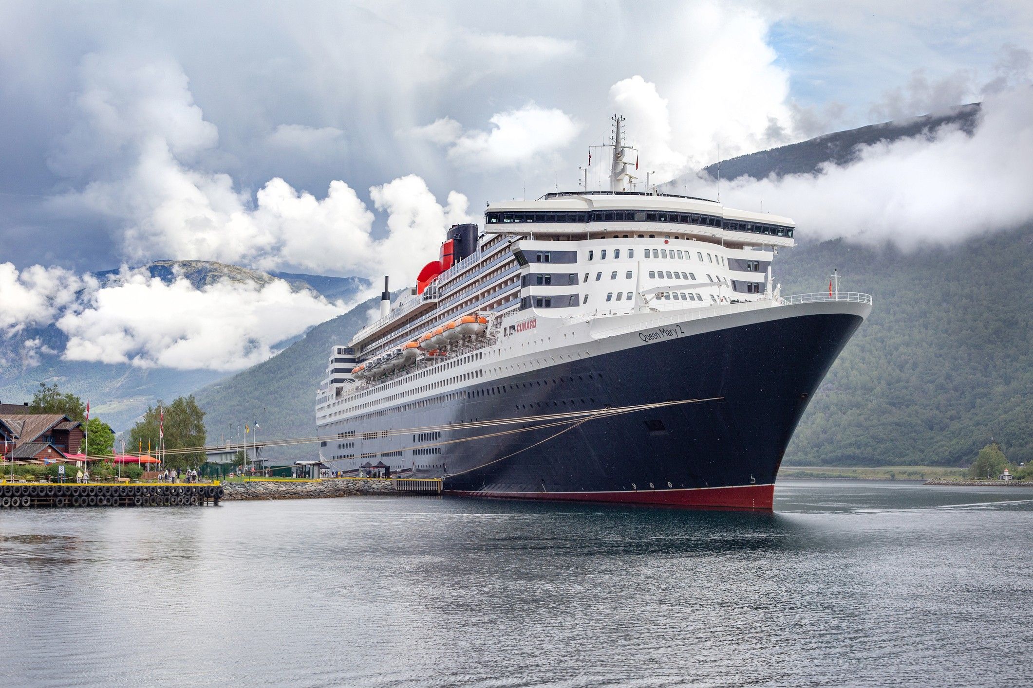 Cunard’s ocean liner and flag ship, Queen Mary 2, docked at the stunning venue of Flam in the Norweigian Fjords surrounded by clouds hanging low over the surrounding mountain landscape. Travelers returning to cruises